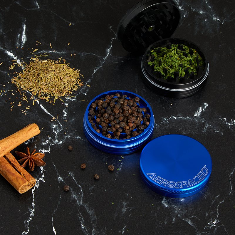 Open blue AEROSPACED grinder filled with peppercorns beside a black grinder holding fresh herbs
